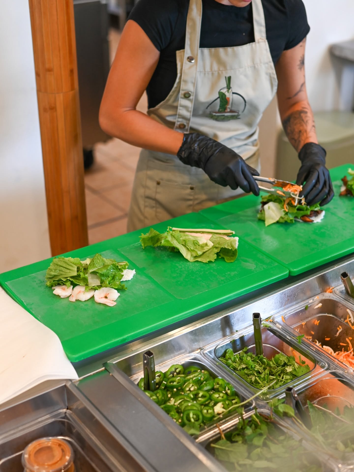 Gloved hands assembling a roll on the green prep board above steel inserts of herbs