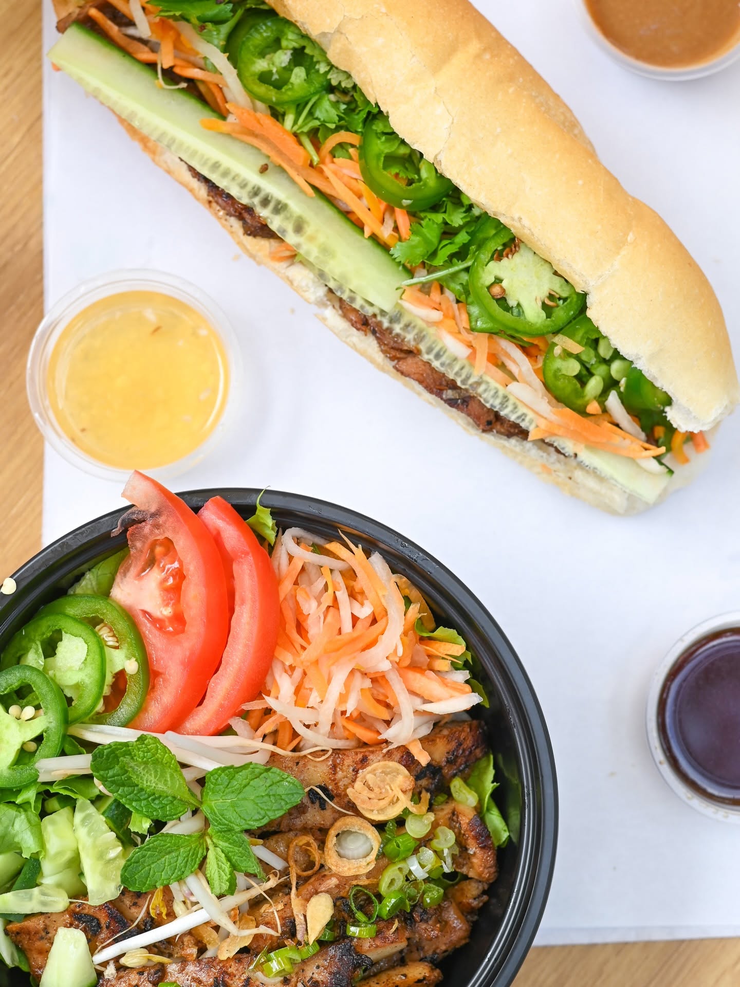 Overhead view of a V-Roll bánh mì next to a grilled-meat rice bowl and house sauce cups