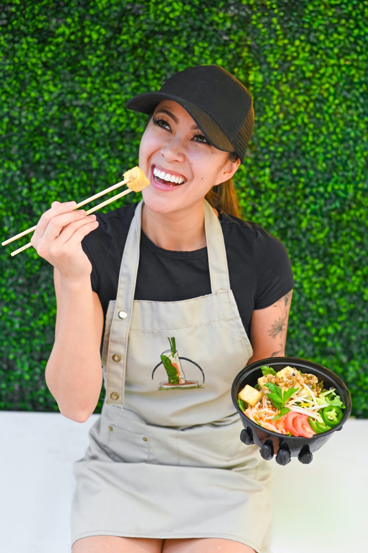 Stephanie Nghiem laughing in a V-Roll apron in front of a green leaf wall, holding a bowl