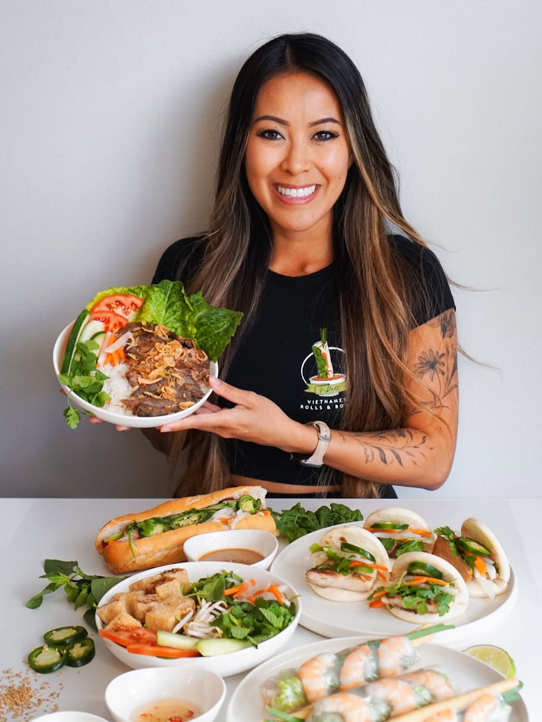 Founder Stephanie Nghiem holding a V-Roll bowl behind a spread of bánh mì, spring rolls and bao